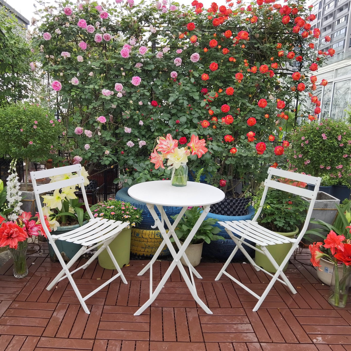 The 3 Piece Patio Bistro Set of a foldable round table and two chairs is placed on a wooden patio, surrounded by green plants and blooming roses, with a vase of flowers on the table.
