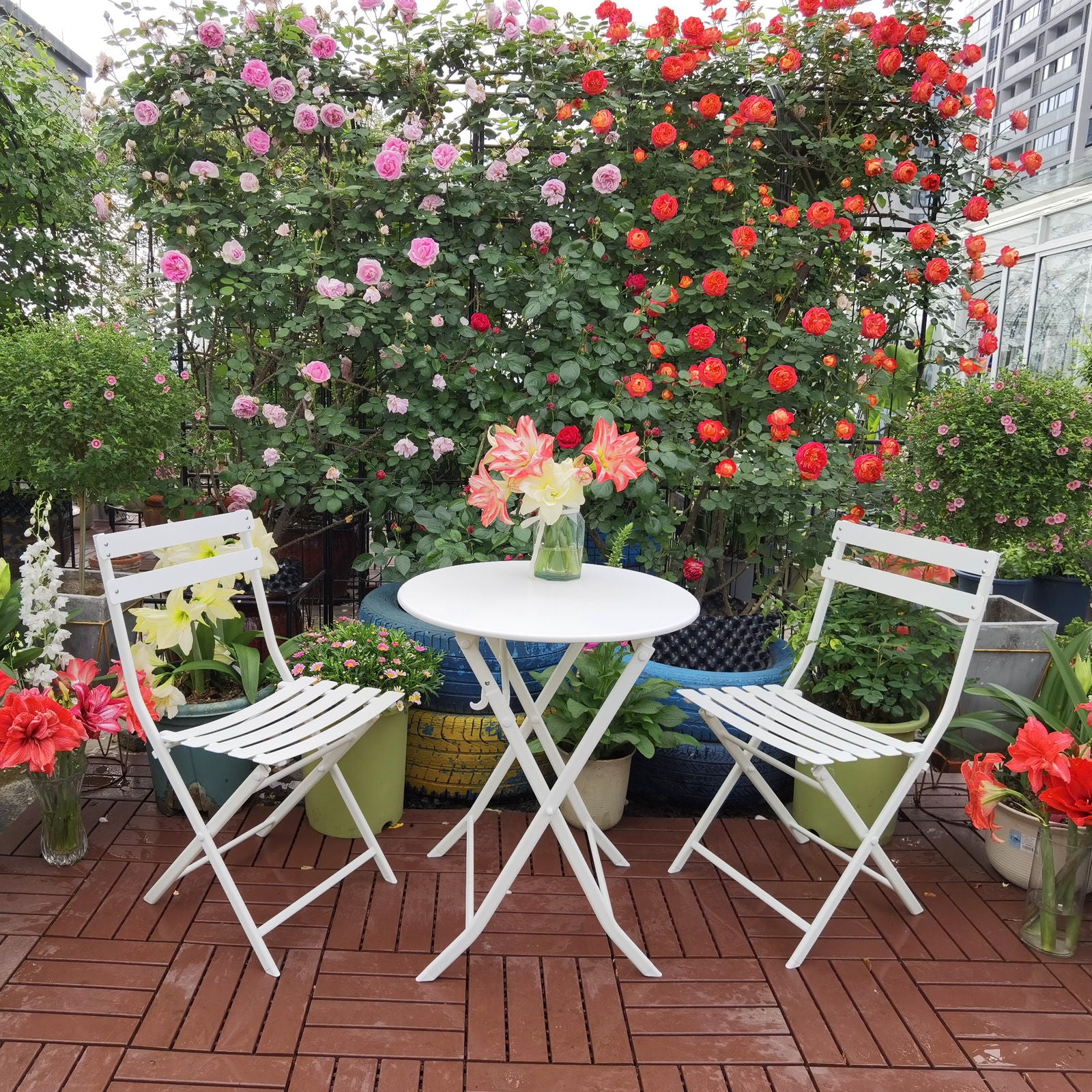 The 3 Piece Patio Bistro Set of a foldable round table and two chairs is placed on a wooden patio, surrounded by green plants and blooming roses, with a vase of flowers on the table.