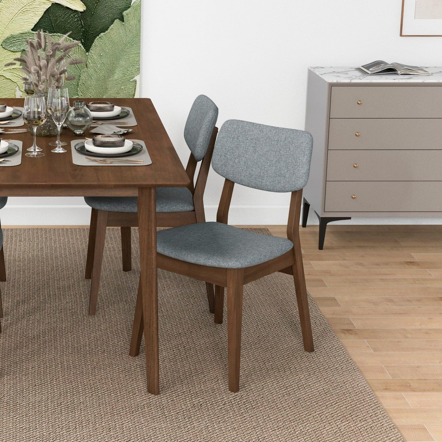 A modern dining room with a wooden table set for four, including Carlos Solid Back Side Chairs in grey, a beige rug, a sideboard with a marble top, and large leafy artwork on the wall.