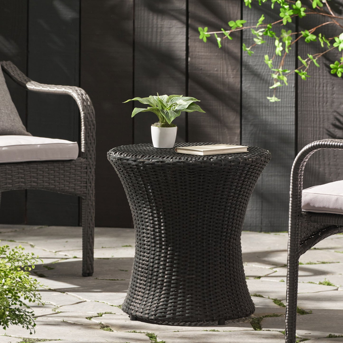 The Thrive Hourglass Rattan Table (no glass top) is placed between two cushioned chairs on a stone patio, styled with a small potted plant and a closed book, surrounded by greenery and set against a black wooden fence.