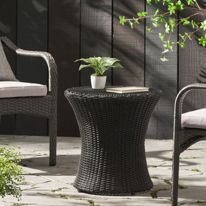 The Thrive Hourglass Rattan Table (no glass top) is placed between two cushioned chairs on a stone patio, styled with a small potted plant and a closed book, surrounded by greenery and set against a black wooden fence.