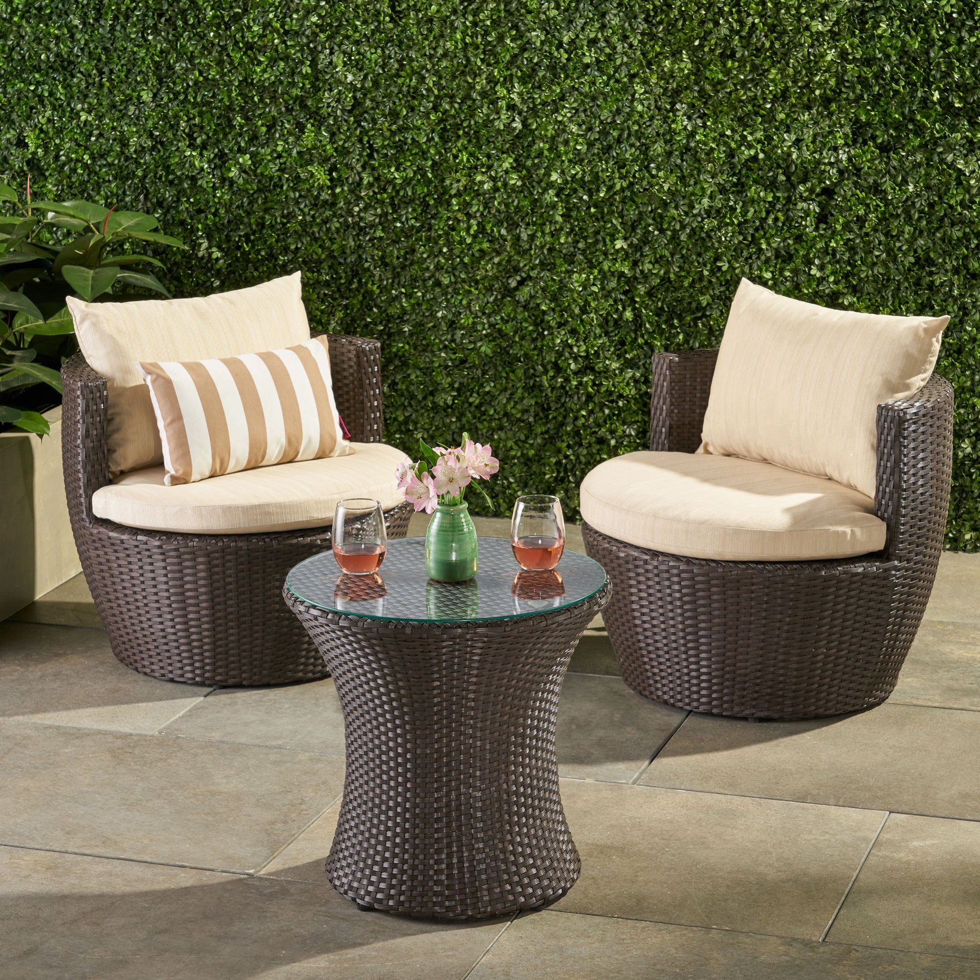 The Kono - Chat Set Outdoor Furniture features two round wicker chairs with cream cushions and a small glass-top table, perfect for your patio against a leafy green backdrop.