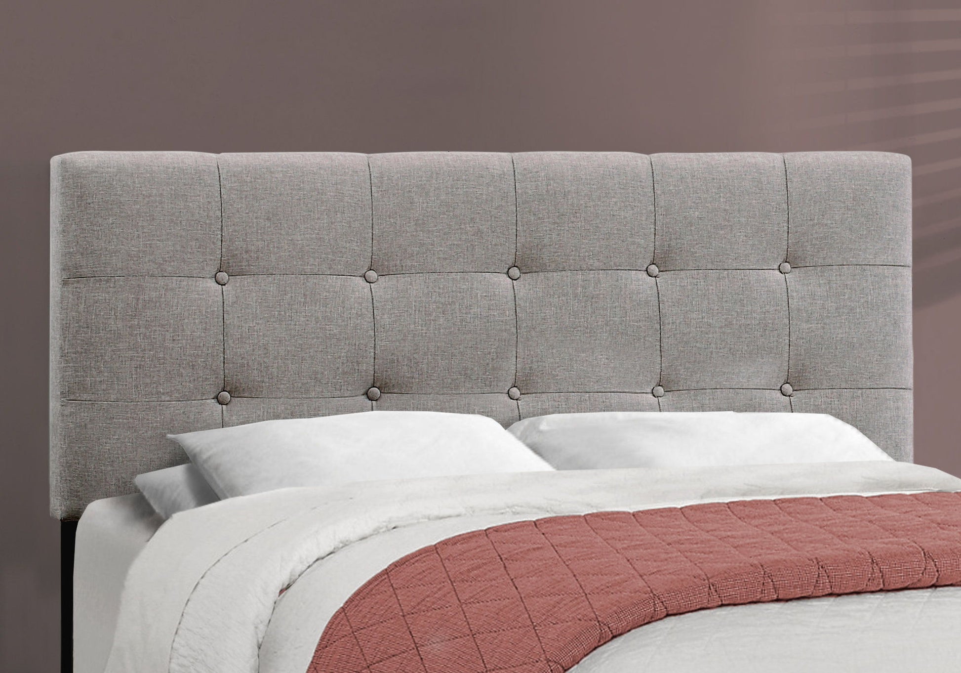 A transitional upholstered bed frame with a light gray tufted headboard, white pillows and bedding, a mauve quilt at the foot, all set against a taupe wall.