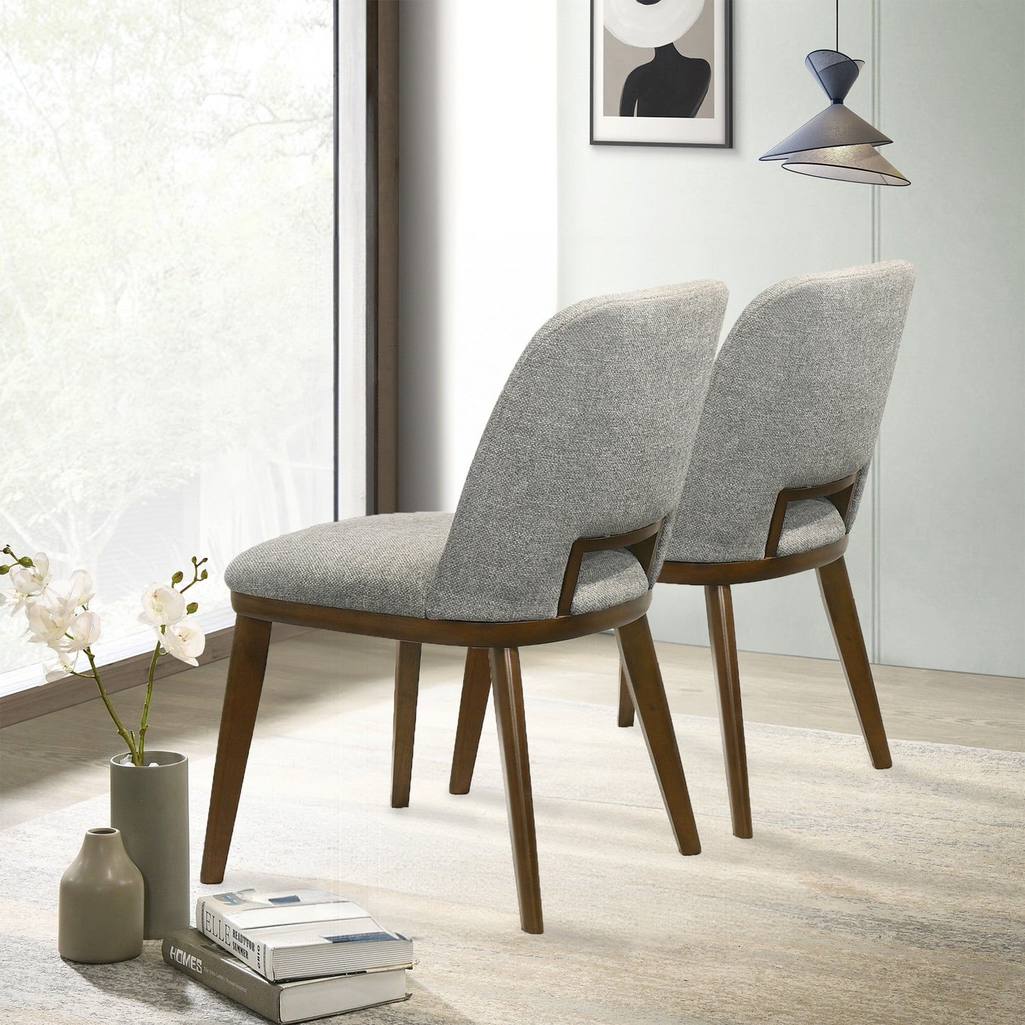 Two Blake - Modern Dining Chairs with light gray upholstered seats, backs, and wooden legs sit side by side on a light rug near a large window. A vase with white flowers and contemporary wall art are in the background.