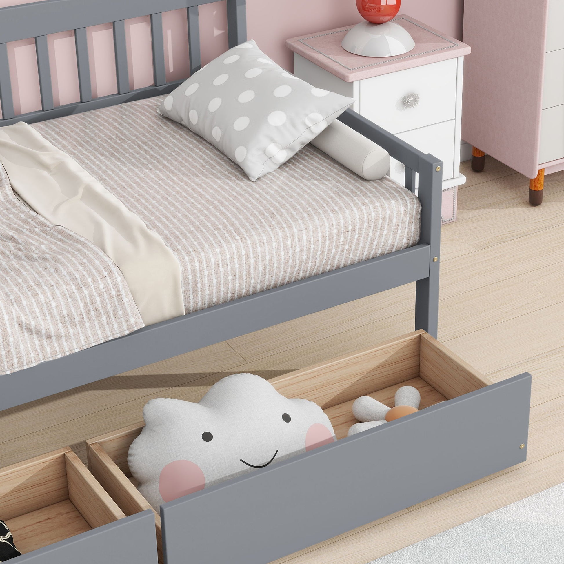 A tidy bedroom features a gray Pine Wood Daybed with two storage drawers, beige-striped bedding, a polka dot pillow, and a white nightstand. Under the daybed, a wooden drawer stores a smiling cloud pillow and a gray stuffed toy.