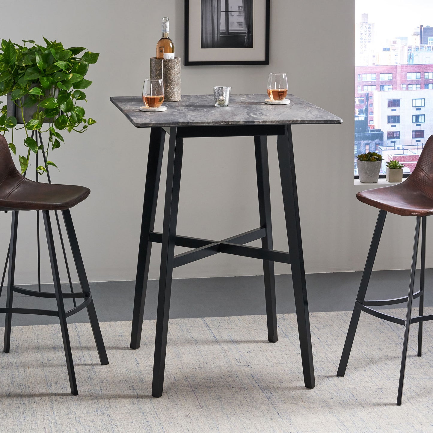 A Modern Bar Height Dining Table with rubberwood legs and a laminate top holds a bottle of rosé, two glasses, and a candle. Two brown barstools sit beside the table, with a plant and city view in the background.