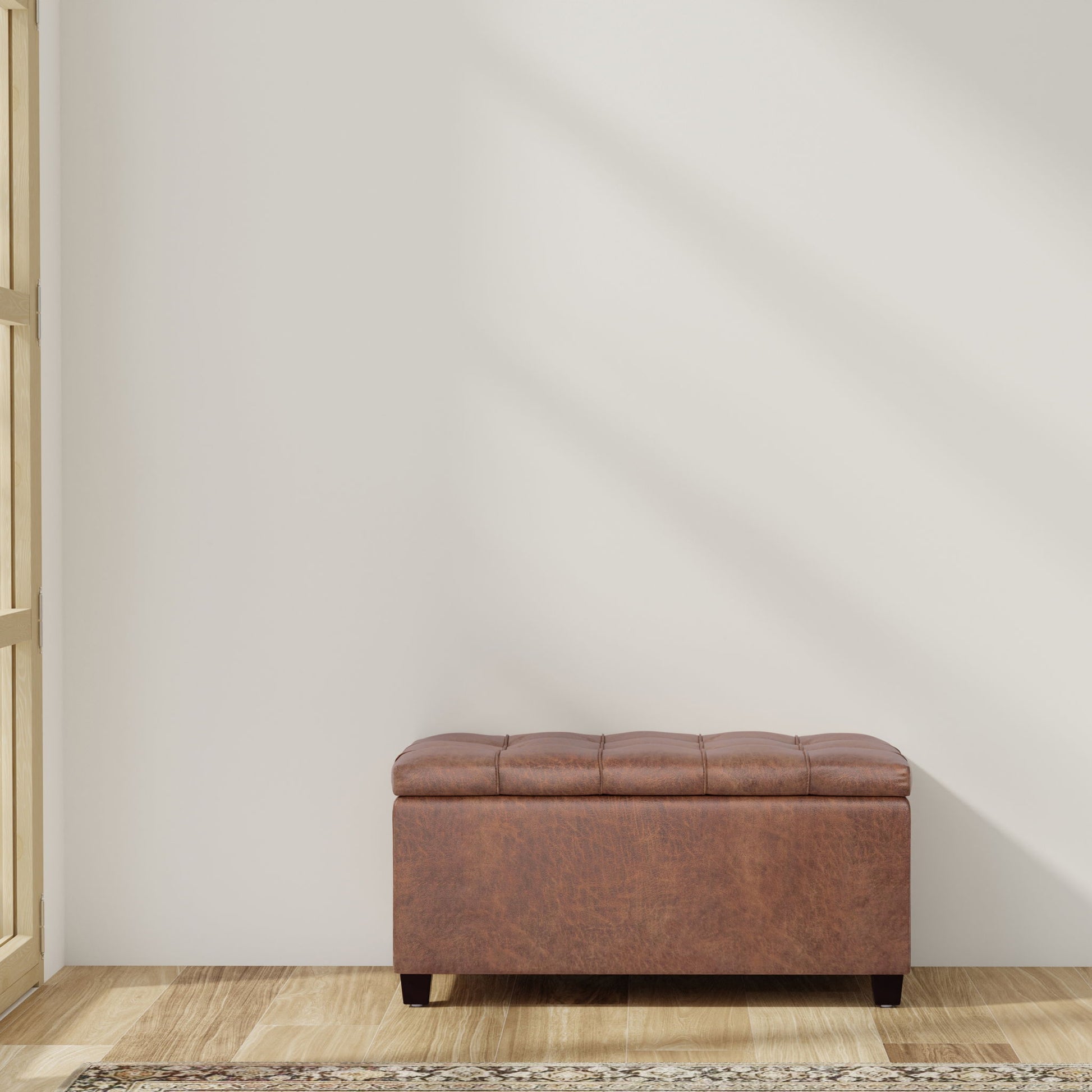 The Sienna - Upholstered Storage Ottoman Bench with a tufted brown leather top sits on wooden flooring against a plain white wall, positioned by a window where sunlight casts soft shadows.