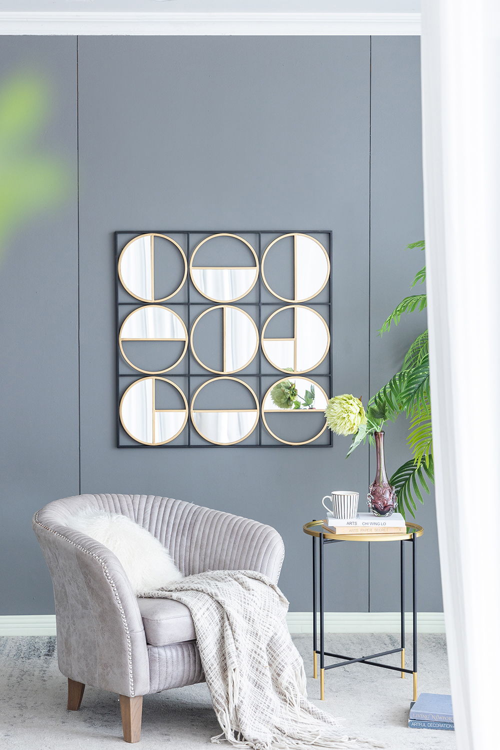 A cozy gray armchair with a white throw blanket sits by a round side table holding books, a mug, and a vase. The gray wall displays the Eclectic Styling Metal Beaded Wall Mirror. Green plants bring freshness to the space.