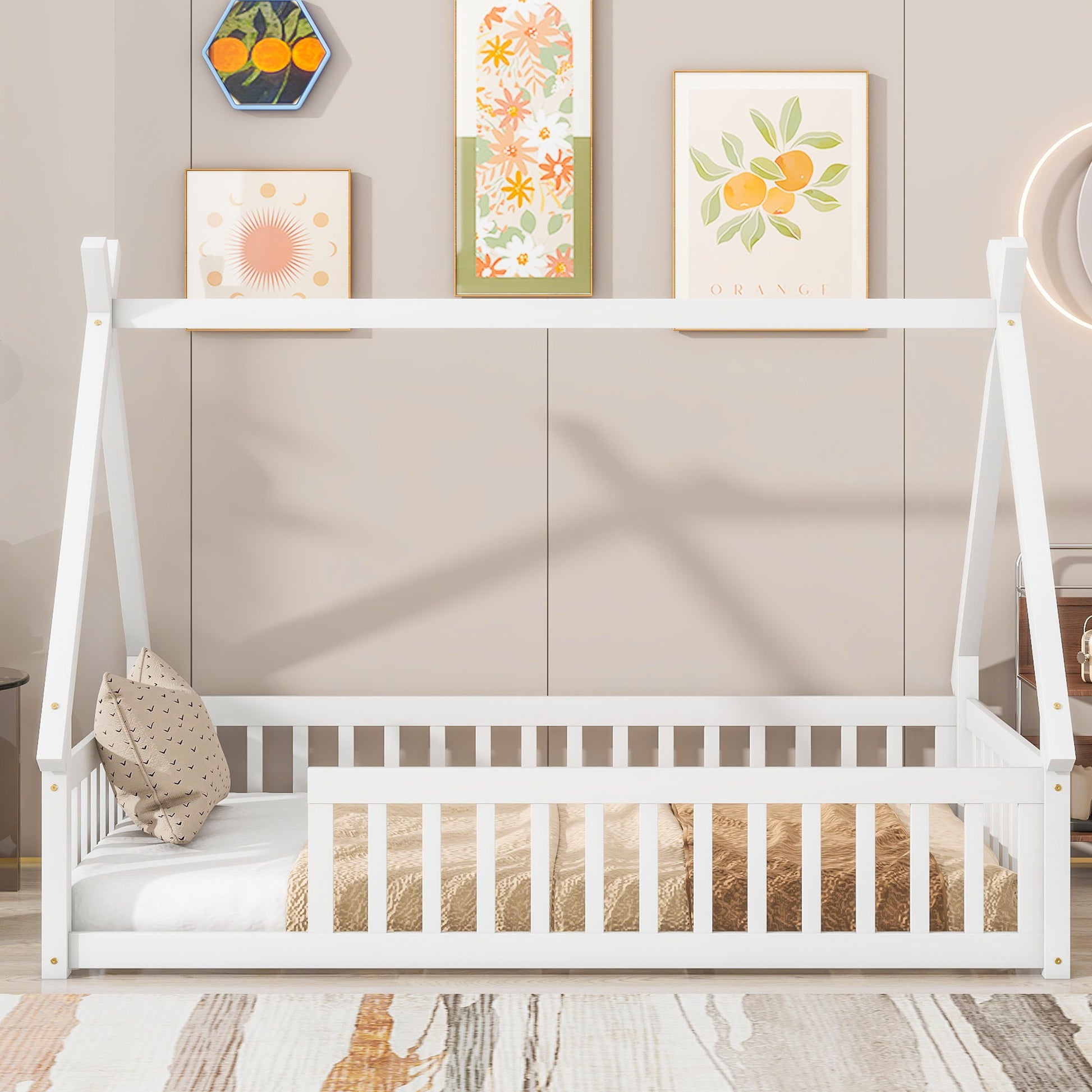 A white Tent-Shaped Floor Bed with guardrails and slats, styled like a teepee, sits against a beige wall with colorful fruit and floral art. Two pillows, a blanket, and a patterned rug complete the cozy toddler room.