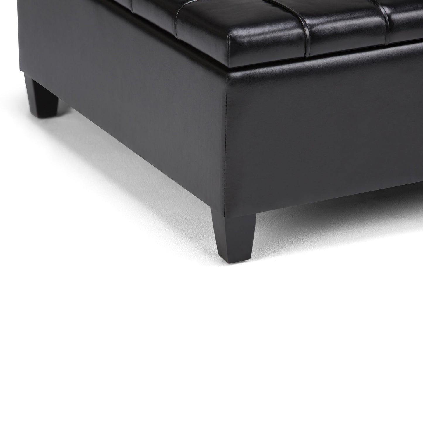 Close-up of the bottom corner of the Harrison - Large Coffee Table Storage Ottoman in black leather with short square wooden legs, placed on a white surface.