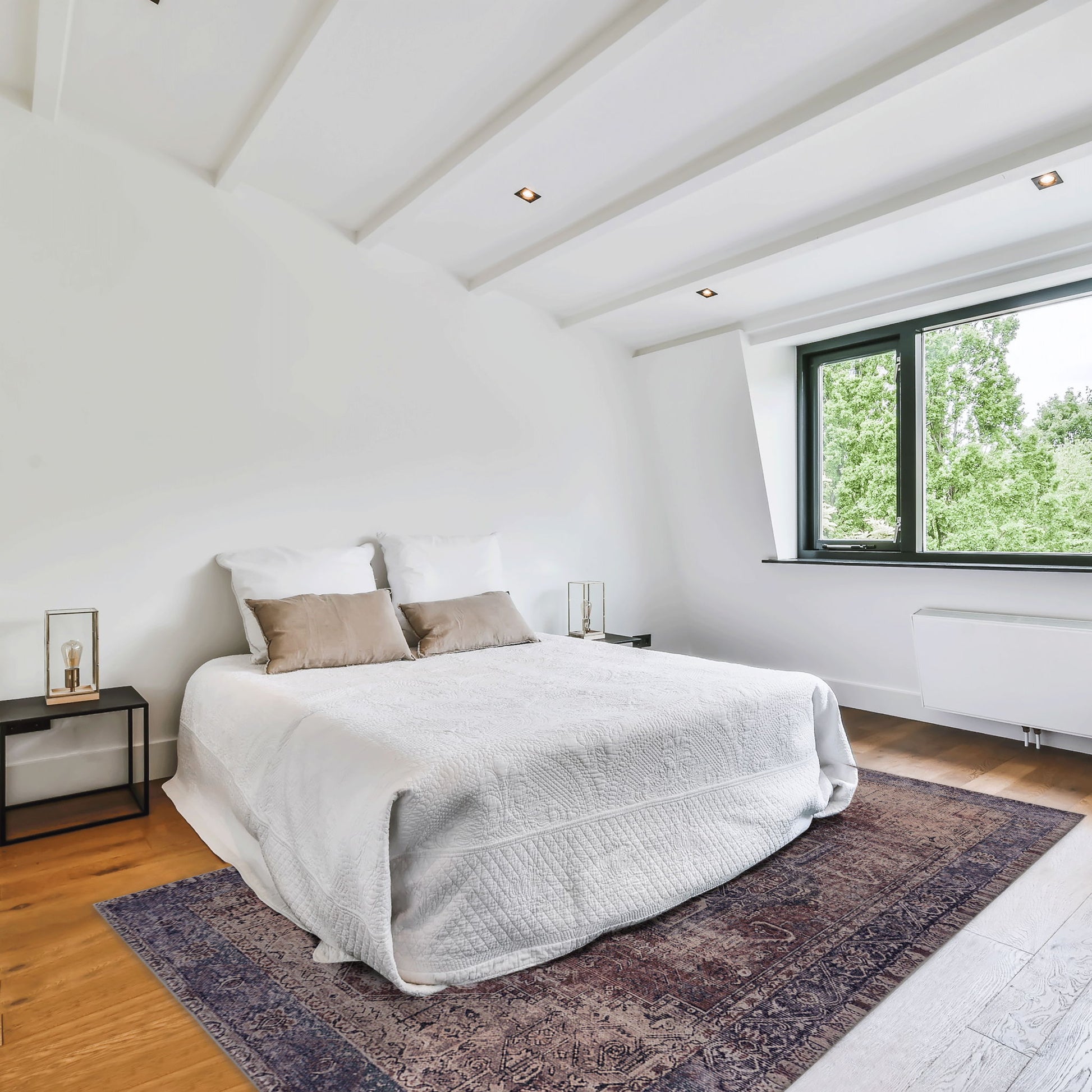 A minimalist bedroom with white walls, a large bed with white bedding and beige pillows, wooden floor, two small bedside tables, a window with a view of greenery, and the Maval - Unique Washable Area Rug.