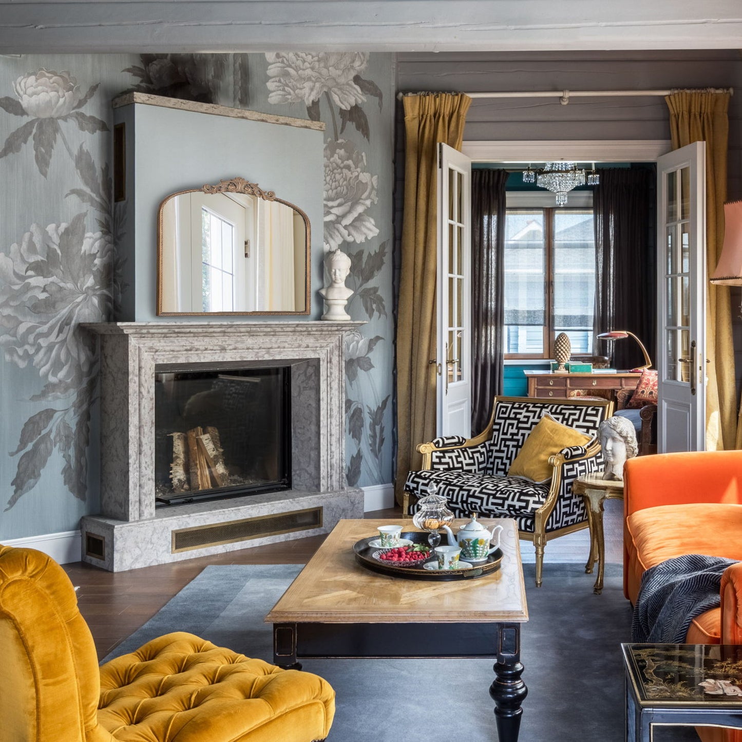 A stylish living room with an Arch Mirror—baroque-inspired wall decor—marble fireplace, floral wallpaper, velvet chairs, wooden coffee table, and chic accents. French doors open to a bright blue-walled space with a chandelier.
