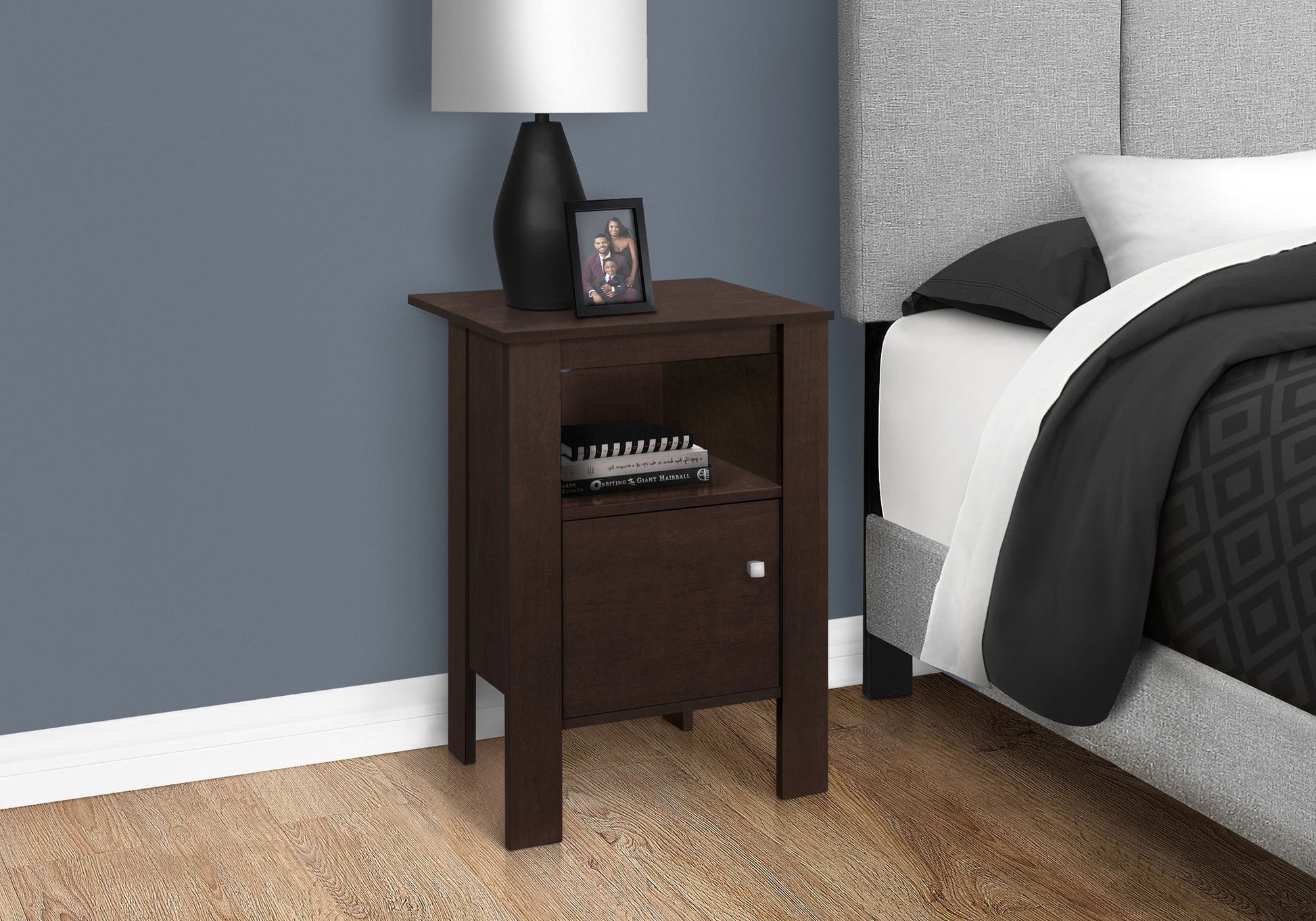 An Accent Nightstand with storage and transitional style sits by the bed with gray and black bedding. A black lamp and framed photo rest on top. The blue wall contrasts with a light wood floor.