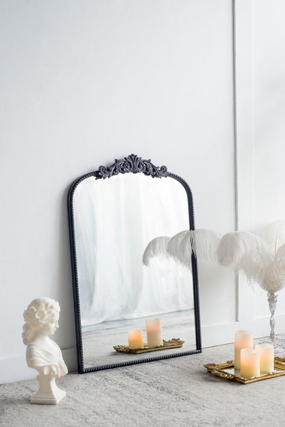 The Arch Mirror, a baroque-inspired wall decor piece, leans against a white wall with a classical bust, gold tray with candles, and a glass vase of white feathers arranged on a soft gray carpet nearby.