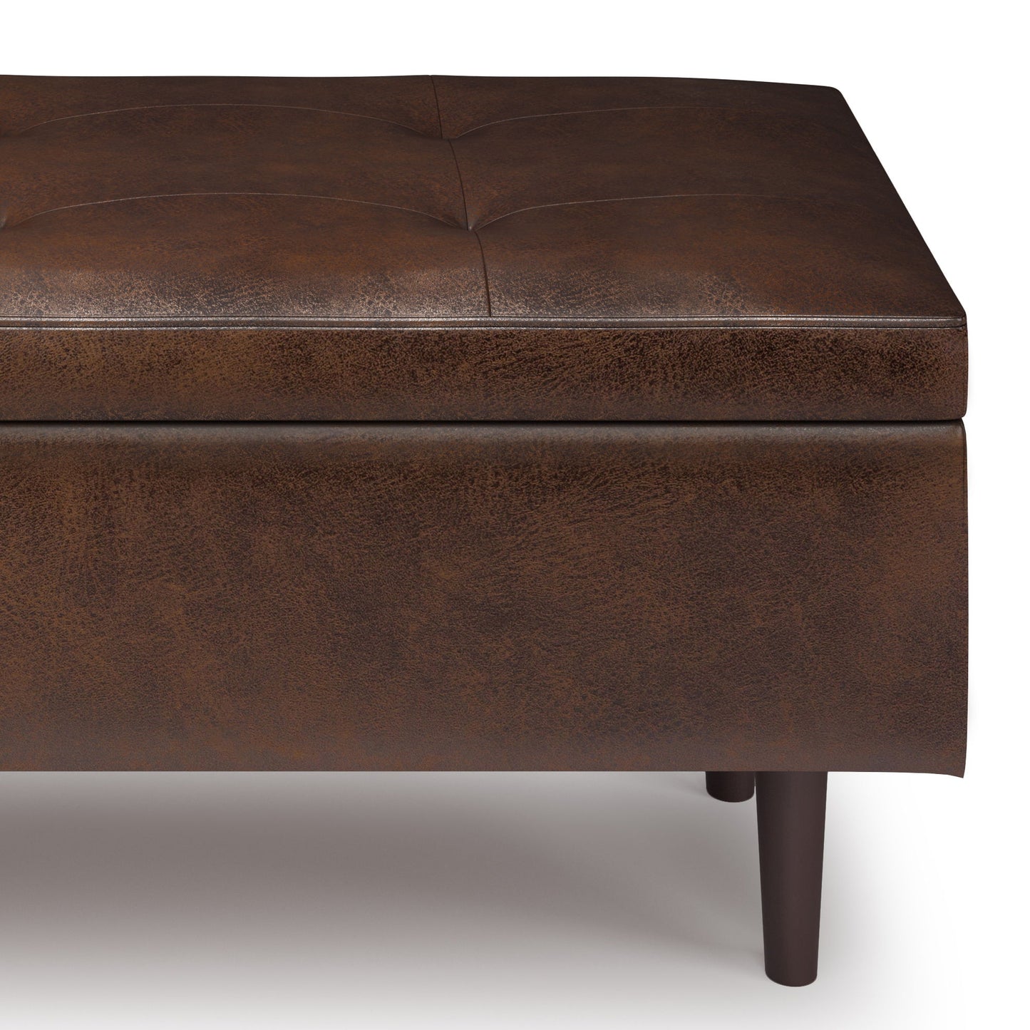 A close-up of the Shay Large Square Coffee Table Storage Ottoman, featuring brown leather upholstery, tufted stitching, and dark wooden legs, shown against a white background.