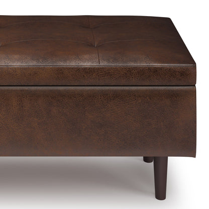 A close-up of the Shay Large Square Coffee Table Storage Ottoman, featuring brown leather upholstery, tufted stitching, and dark wooden legs, shown against a white background.