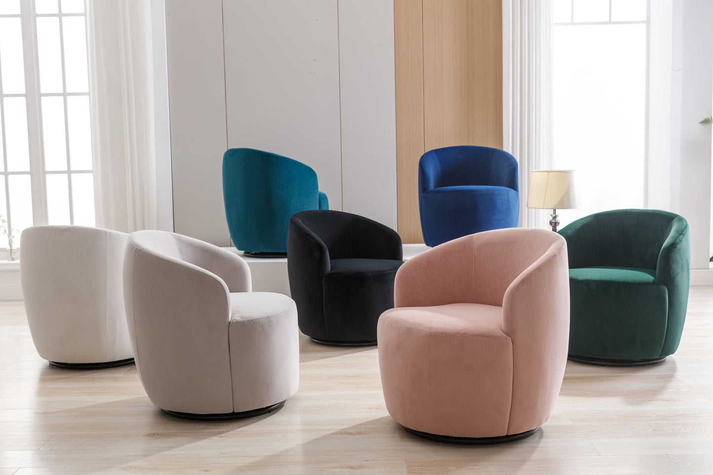 Six Velvet Fabric Swivel Accent Armchair Barrel Chairs with powder-coated metal rings in pastel pink, white, dark green, teal, black, and blue are arranged on a light wood floor in a bright room with large windows and white walls.