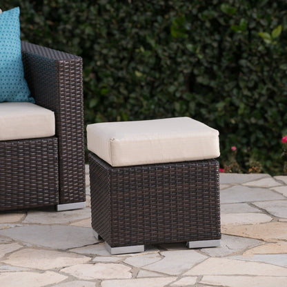 The Santa Rosa Outdoor Lounge Ottoman, featuring dark brown wicker and a light beige cushion, sits on a stone patio beside a matching chair with blue pillows and green foliage in the background.