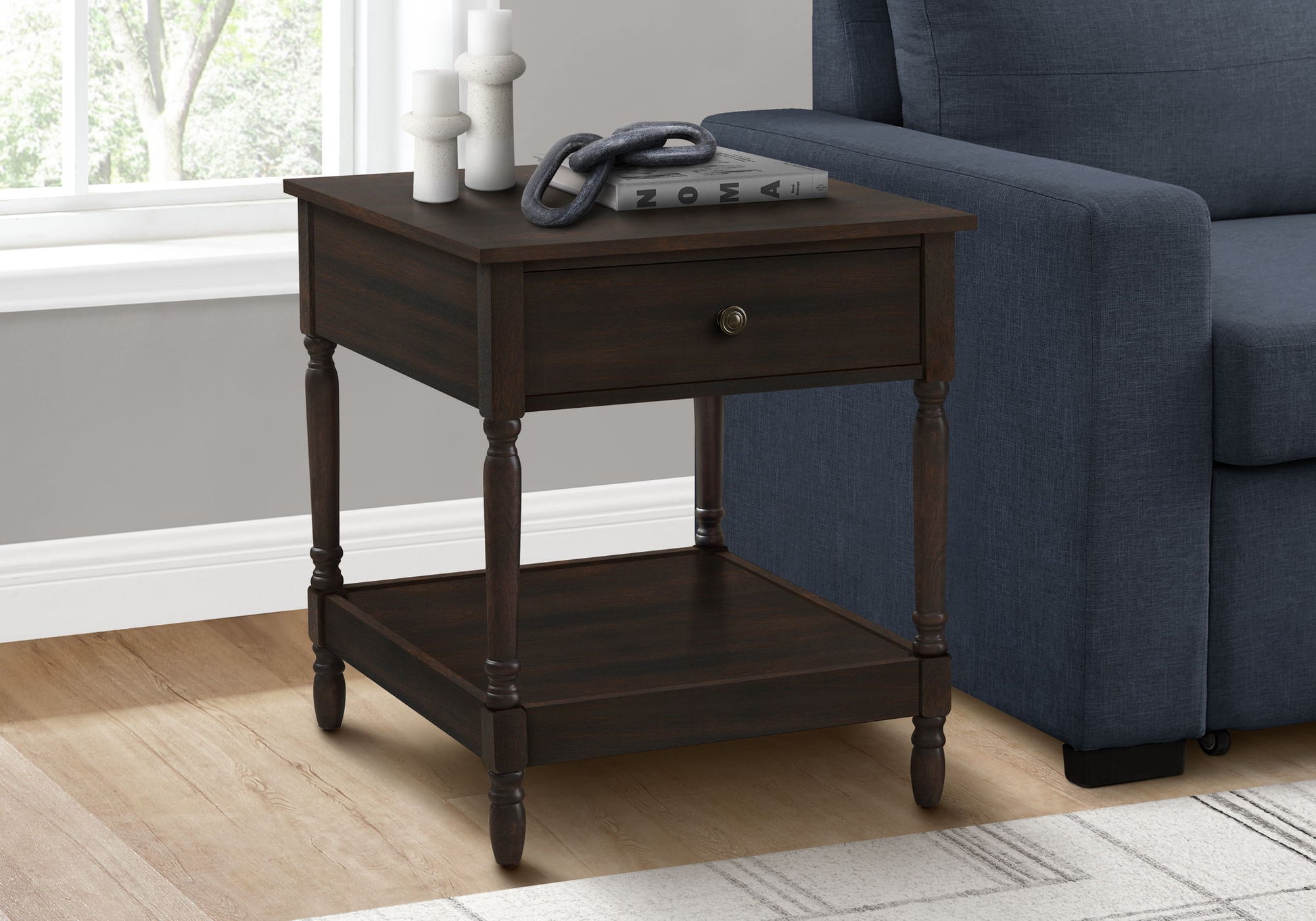 The Accent - Table, 2 Tier, Square, Traditional - Walnut sits by a blue sofa on a light wood floor. Sunlight streams in as books, decor pieces, and candles rest on its surface and lower shelf.