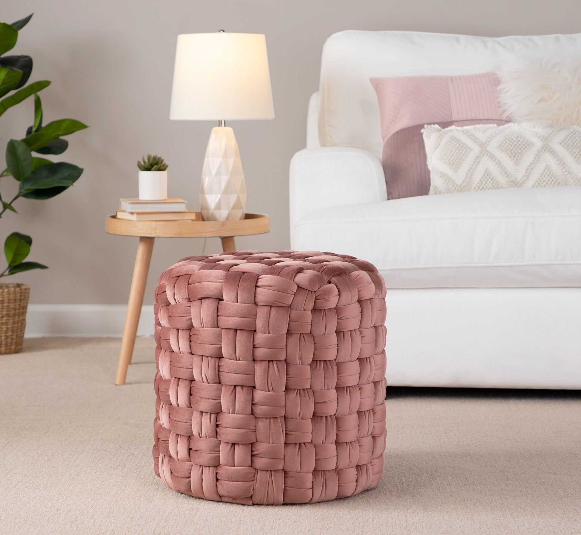 A Braided - Versatile Round Ottoman in pink sits on a beige carpet in a living room featuring a white sofa, decorative pillows, a small side table with a lamp, and a potted plant in the background.