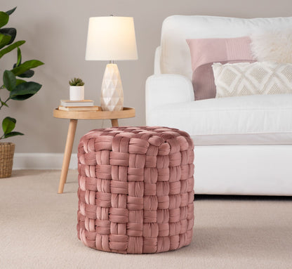 A Braided - Versatile Round Ottoman in pink sits on a beige carpet in a living room featuring a white sofa, decorative pillows, a small side table with a lamp, and a potted plant in the background.