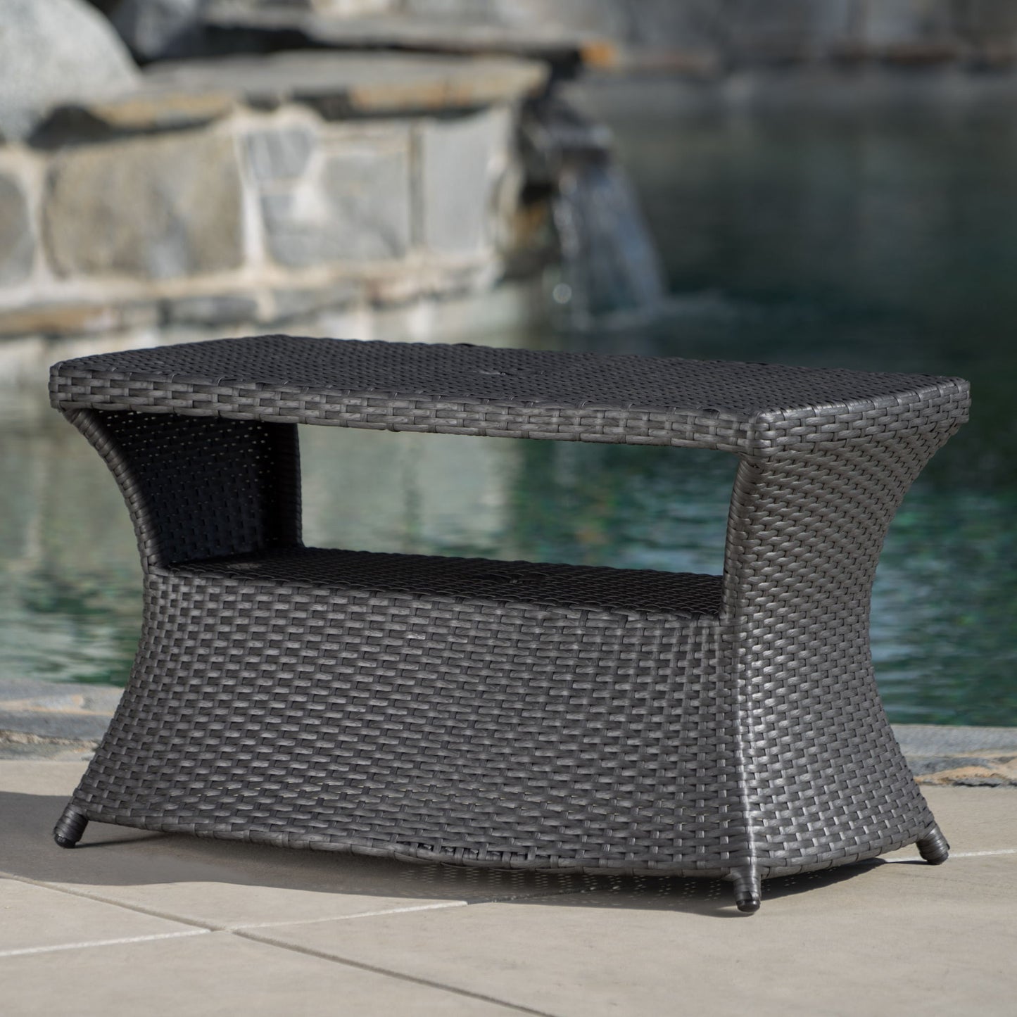 The Berkeley - Chaise Lounge Side Table is a dark brown wicker outdoor table with a lower shelf, shown on a patio near a pool featuring stonework and a waterfall in the background.