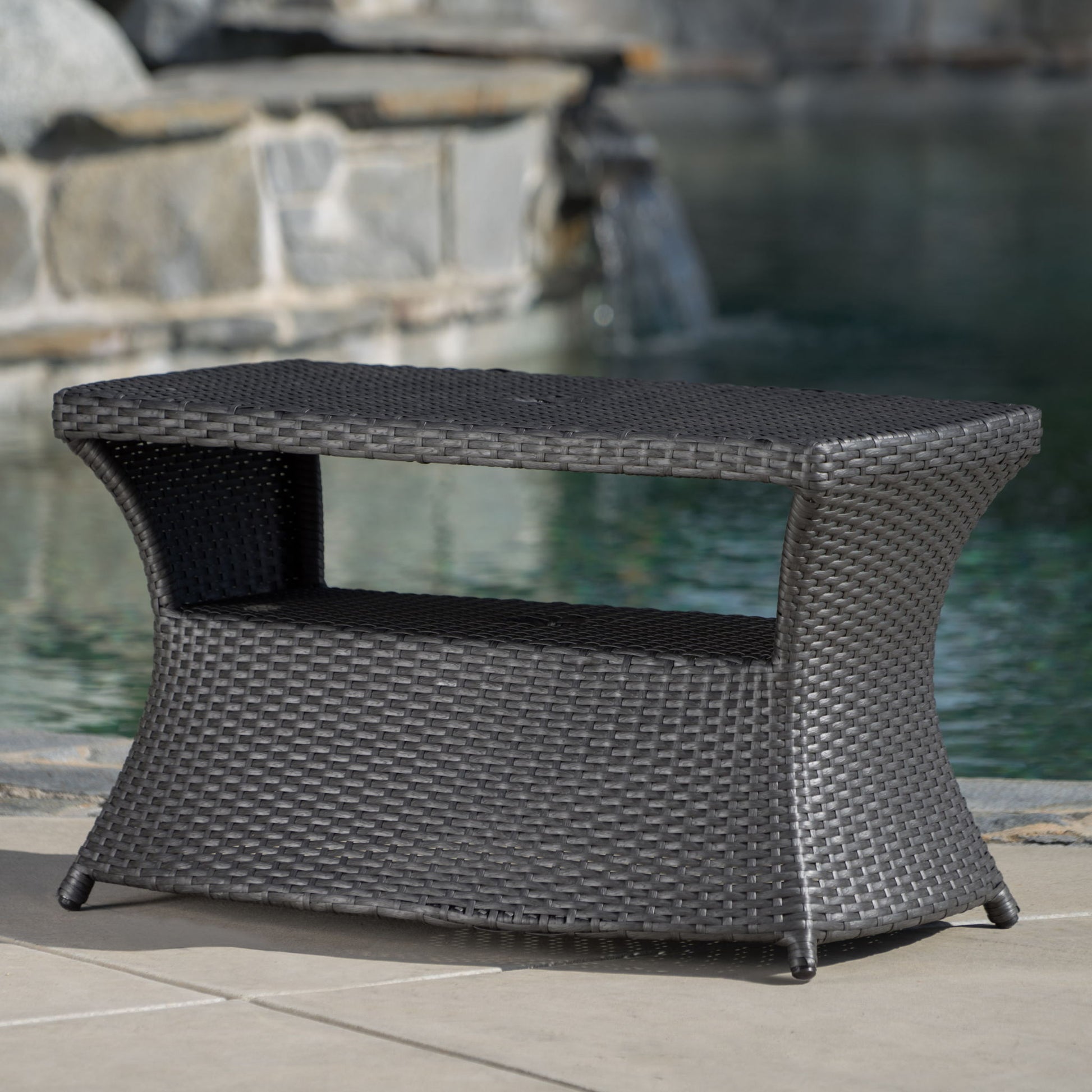 The Berkeley - Chaise Lounge Side Table is a dark brown wicker outdoor table with a lower shelf, shown on a patio near a pool featuring stonework and a waterfall in the background.