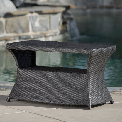 The Berkeley - Chaise Lounge Side Table is a dark brown wicker outdoor table with a lower shelf, shown on a patio near a pool featuring stonework and a waterfall in the background.