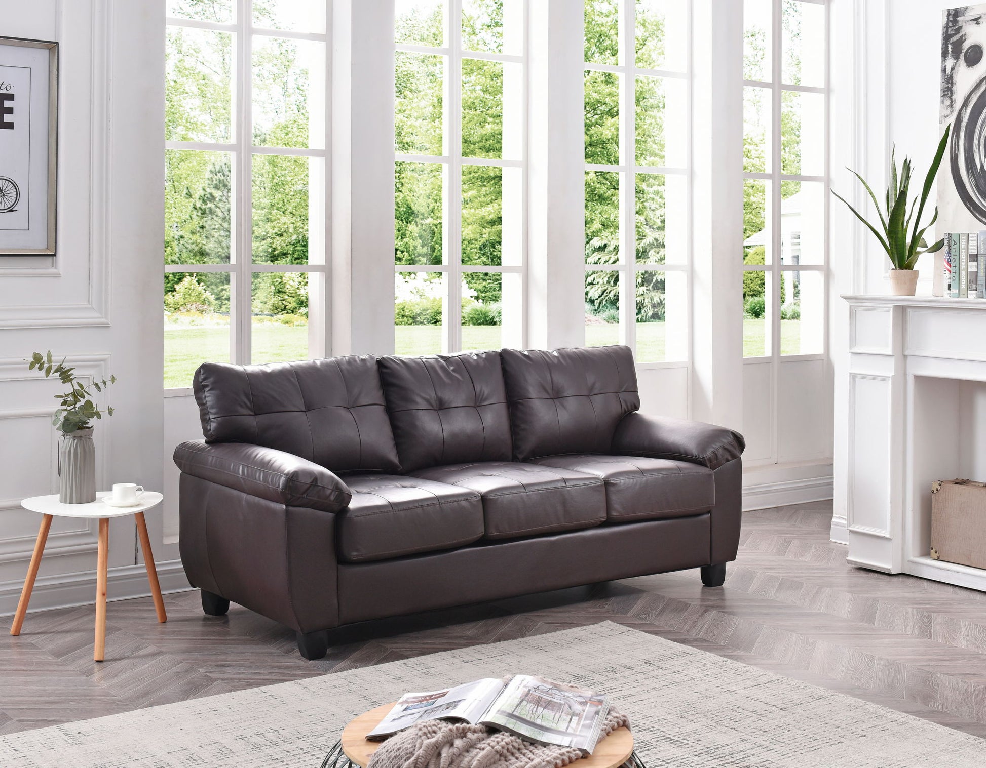 The Comfortable Sofa, in dark brown leather, is featured in a bright living room with large windows and modern decor. A small side table displays a cup and vase, while a round coffee table in front holds magazines.
