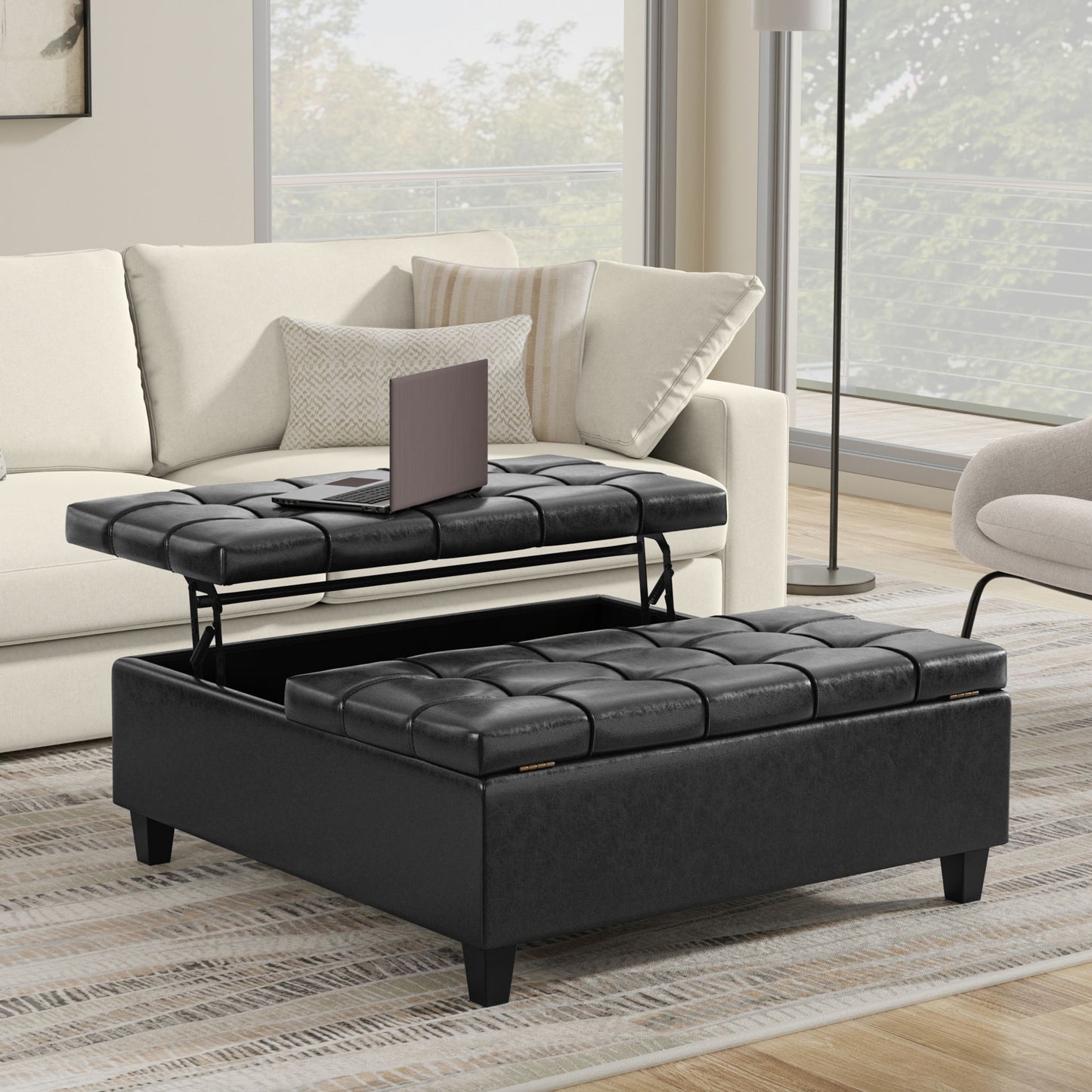 The Harrison Upholstered Coffee Table Storage Ottoman, featuring a black tufted design and lift-top tray with storage, sits before a beige sofa in a modern living room. A laptop is on its raised tray, illuminated by natural light from large windows.