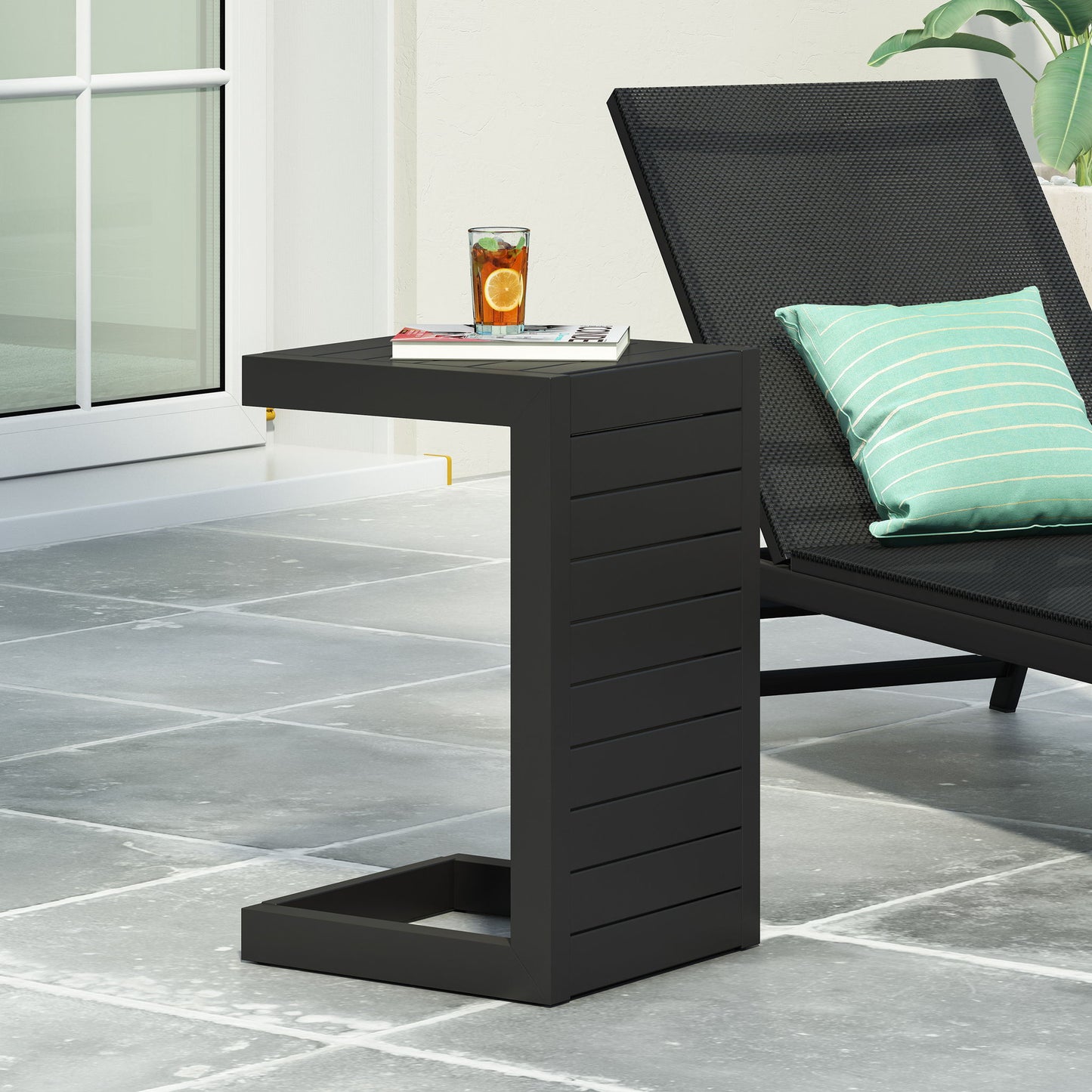 The Aluminum Outdoor C-Shaped Side Table for Patio is placed beside a black lounge chair with a green striped pillow, holding a citrus drink and magazine on a gray tiled patio near a glass door.