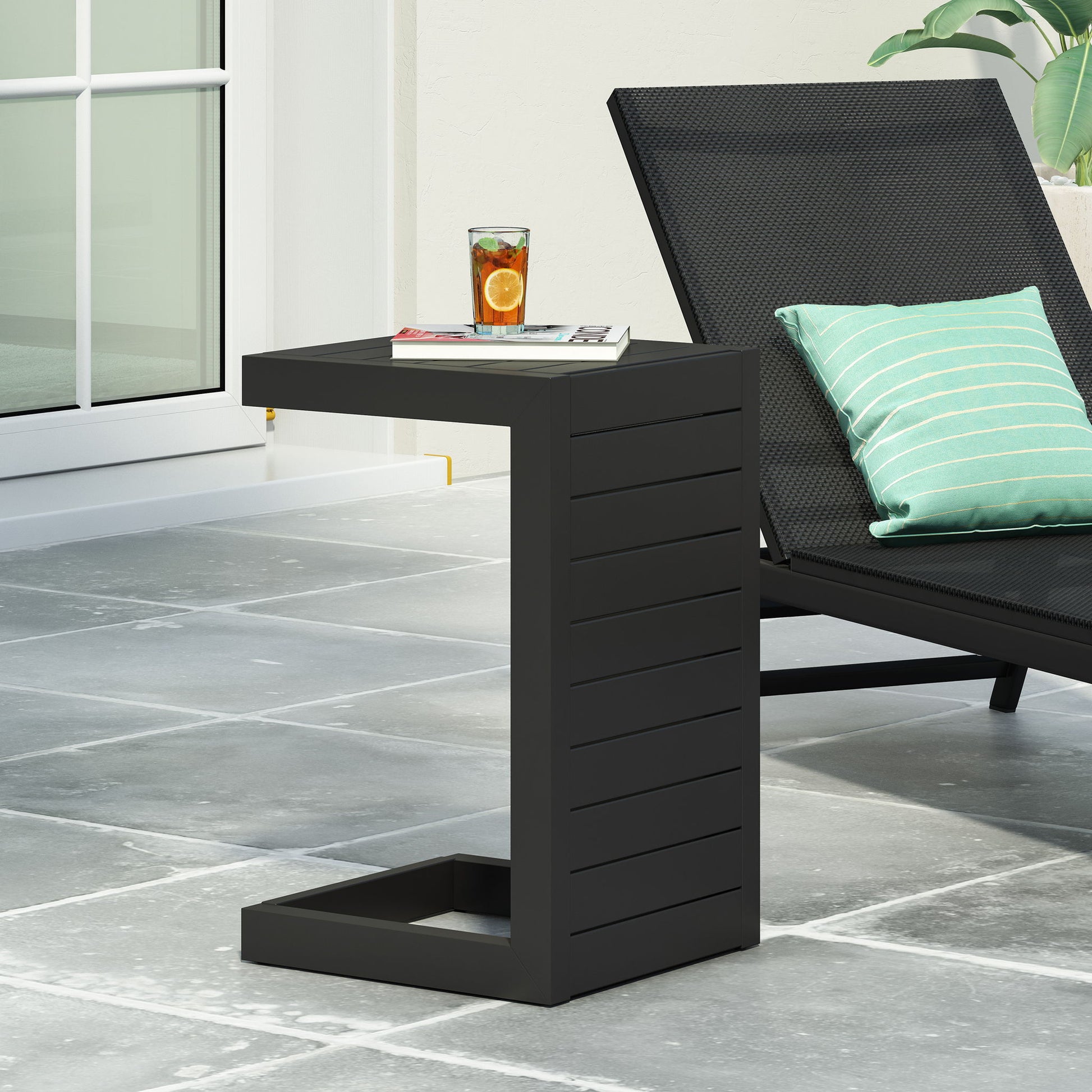 The Aluminum Outdoor C-Shaped Side Table for Patio is placed beside a black lounge chair with a green striped pillow, holding a citrus drink and magazine on a gray tiled patio near a glass door.