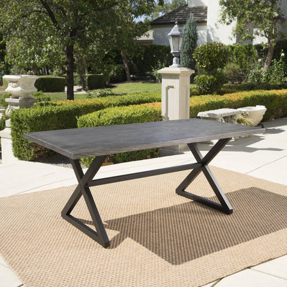 The Rolando Outdoor Aluminum Dining Table with a dark rectangular top and X-shaped black steel legs sits on a tan woven rug in a landscaped patio surrounded by trees, bushes, and garden decor.