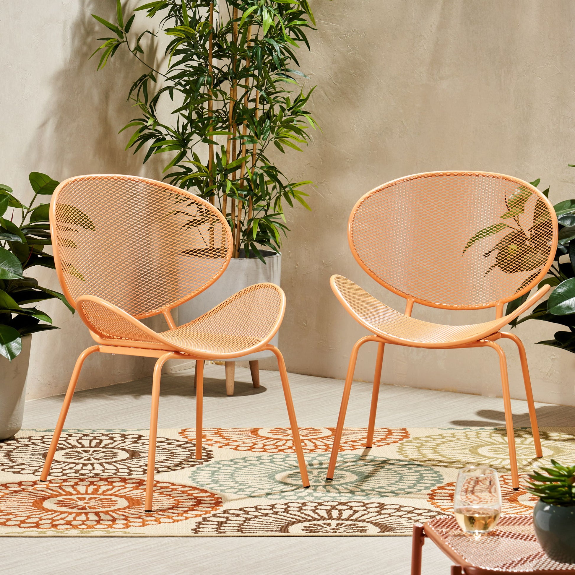 Two Elba Contemporary Chairs for outdoor relaxation, featuring peach-colored metal frames with mesh backs and seats, are placed on a patterned rug amid potted plants. A glass of white wine sits on a table nearby.
