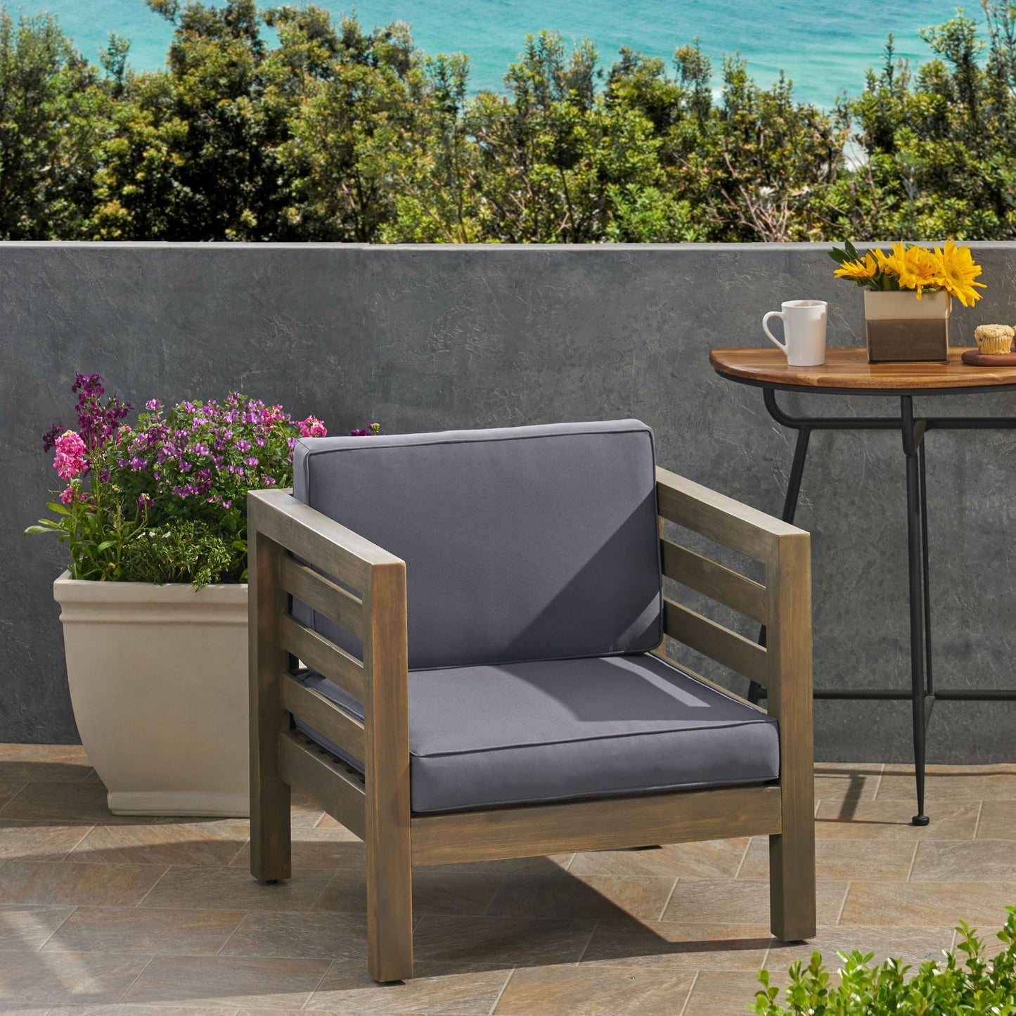 An Outdoor Acacia Wood Patio Chair With Fabric Cushion rests on tiled flooring beside a side table holding coffee and flowers. A large planter with purple blooms is behind, and the ocean can be seen beyond a gray wall.