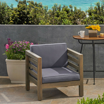 An Outdoor Acacia Wood Patio Chair With Fabric Cushion rests on tiled flooring beside a side table holding coffee and flowers. A large planter with purple blooms is behind, and the ocean can be seen beyond a gray wall.
