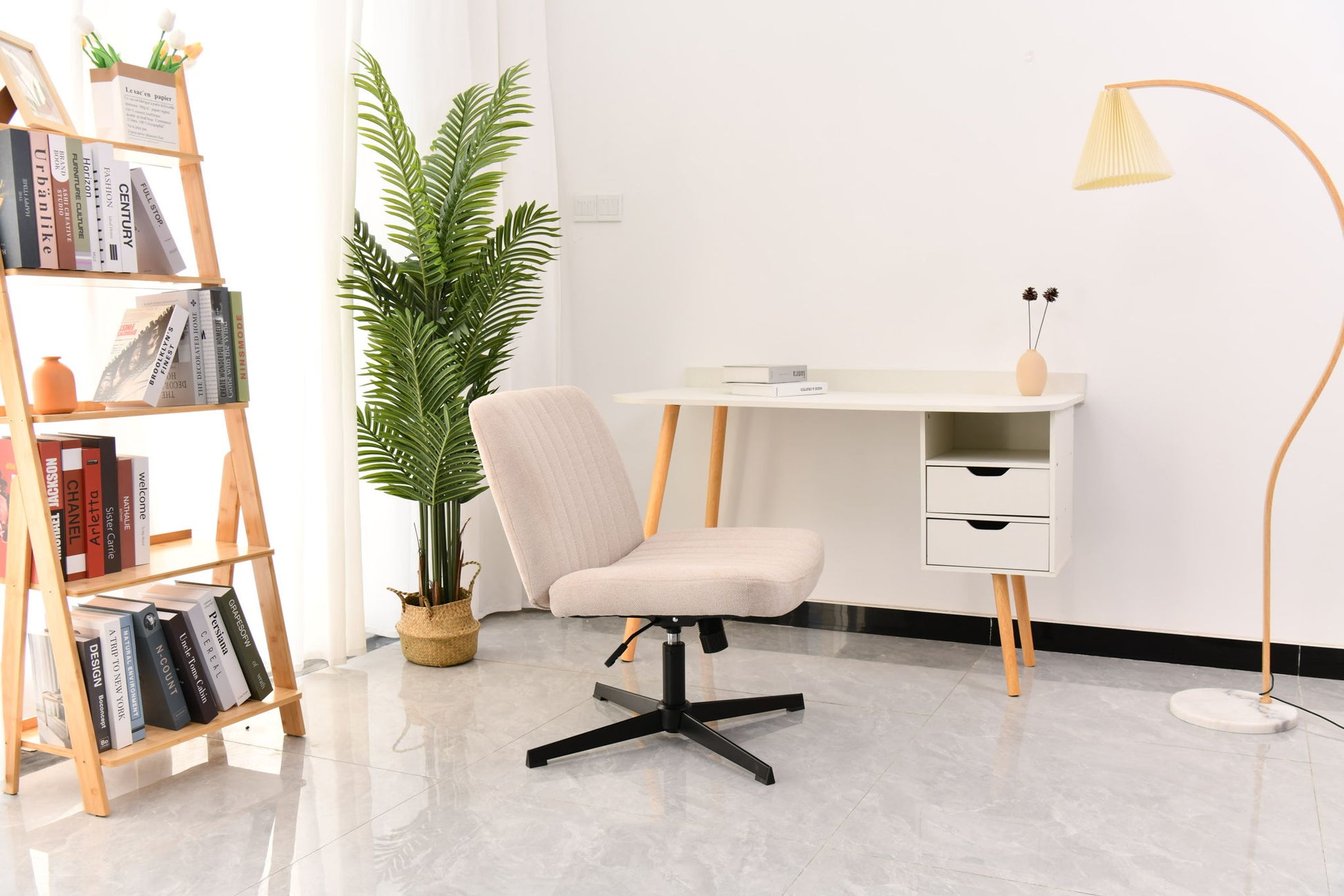 A modern, minimal home office with a beige Cross Legged Office Chair (swivel, height-adjustable), white desk with drawers, a bookshelf full of books, potted plant, floor lamp, and light gray tiled floor. Natural light streams in from the left.