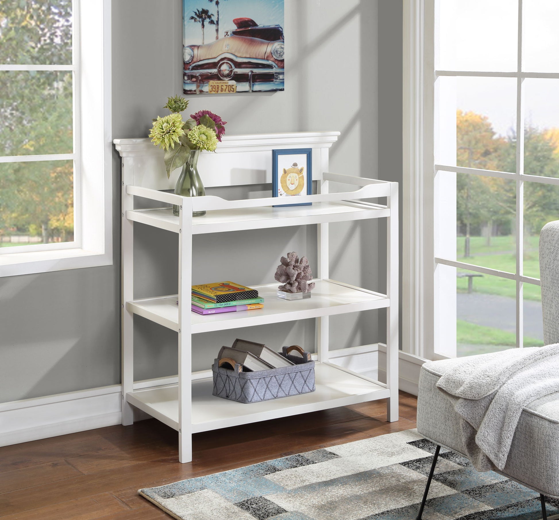 A Universal Changing Table with three white tiers sits by large windows in a bright room. The top tier holds flowers, a photo frame, and books; decor and magazines fill the middle, with a gray basket on the bottom shelf.