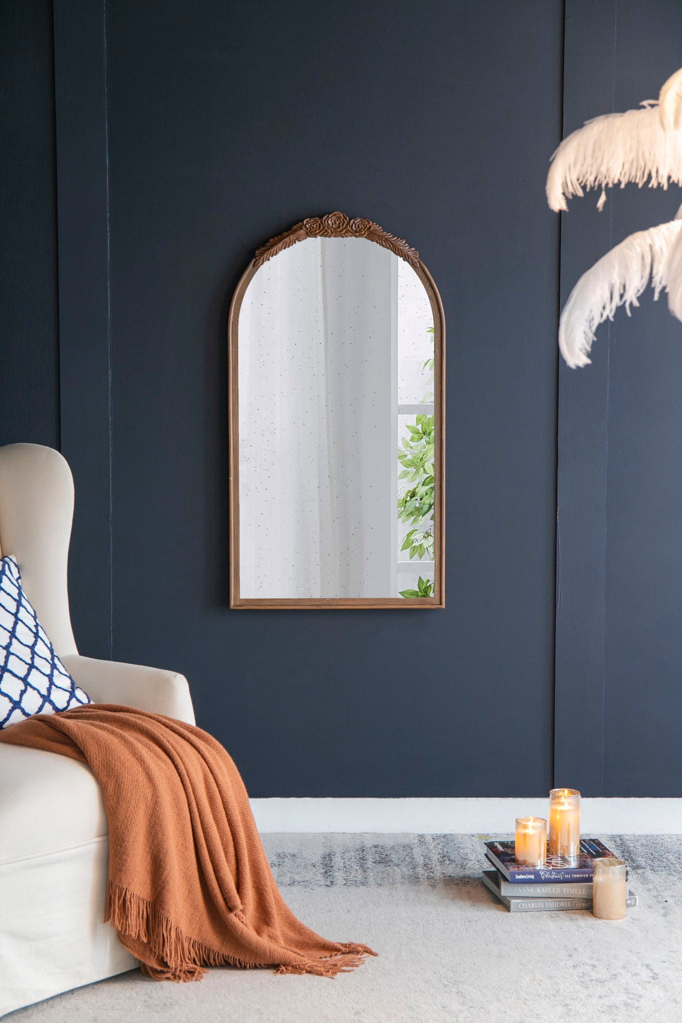 A cozy living room corner with a cream armchair, orange throw, patterned pillow, and a Hand Carved Rose Antique Mirror Frame (Wood Arch Mirror Wall Decor) on a dark blue wall. Candles, books, and white feathered decor rest nearby.