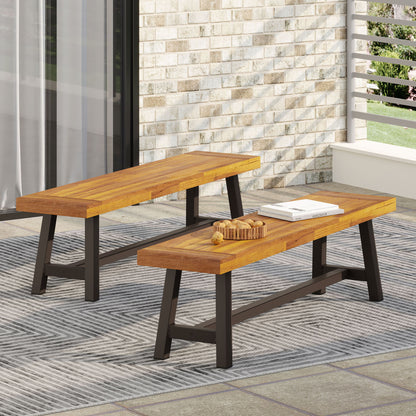 Two Tranquilizer Outdoor Acacia Wood Benches with slat panel design and black metal legs sit on a patterned gray rug. One bench displays a tray with snacks and a book on a patio with brick wall and large glass doors.
