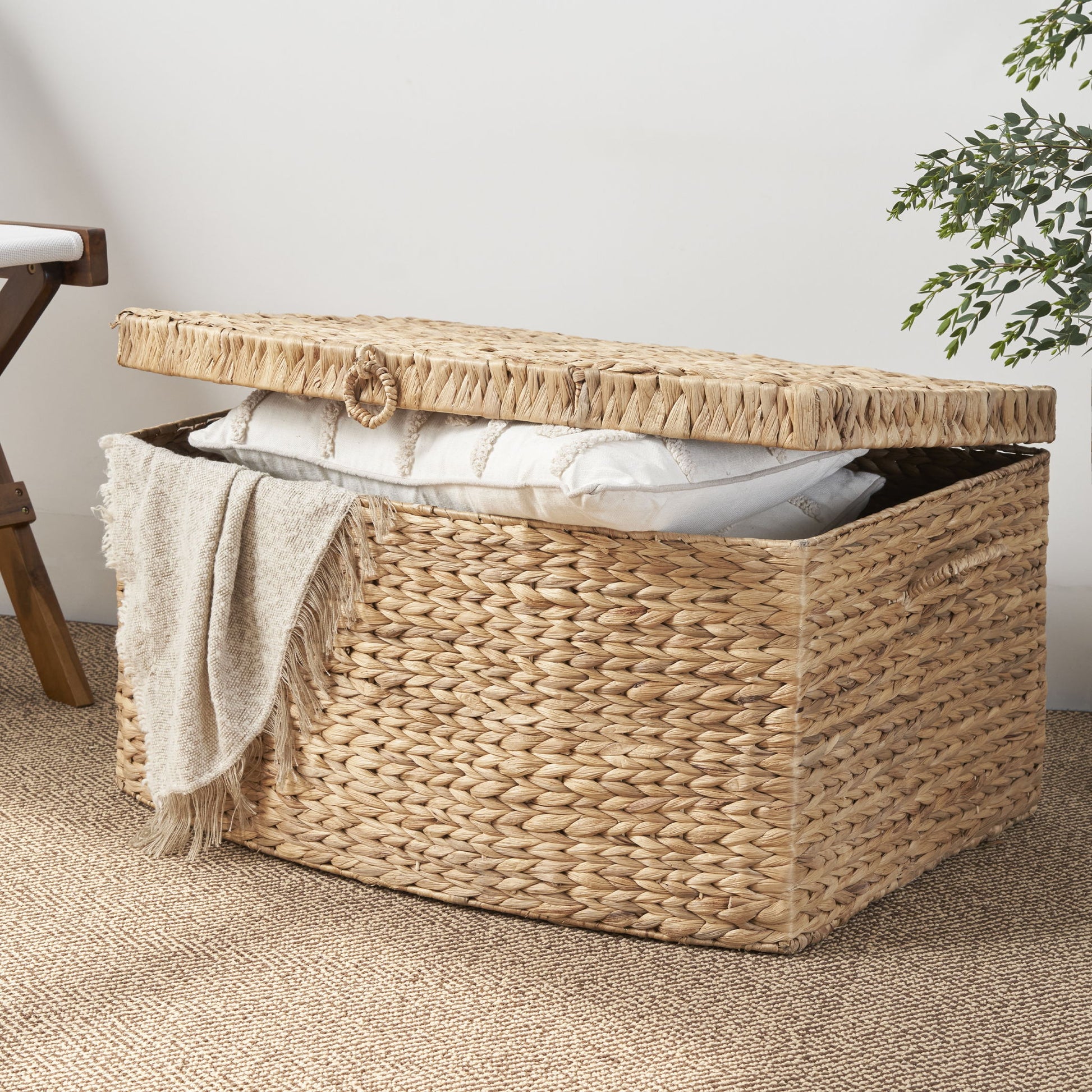The Rectangular Curve Water Hyacinth Woven Wicker Trunk with Handles in natural/light brown sits on a textured rug, its partially open lid revealing a folded blanket and white pillow beside a wooden chair and green plant.