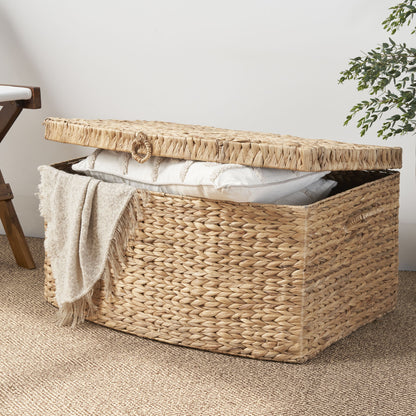 The Rectangular Curve Water Hyacinth Woven Wicker Trunk with Handles in natural/light brown sits on a textured rug, its partially open lid revealing a folded blanket and white pillow beside a wooden chair and green plant.