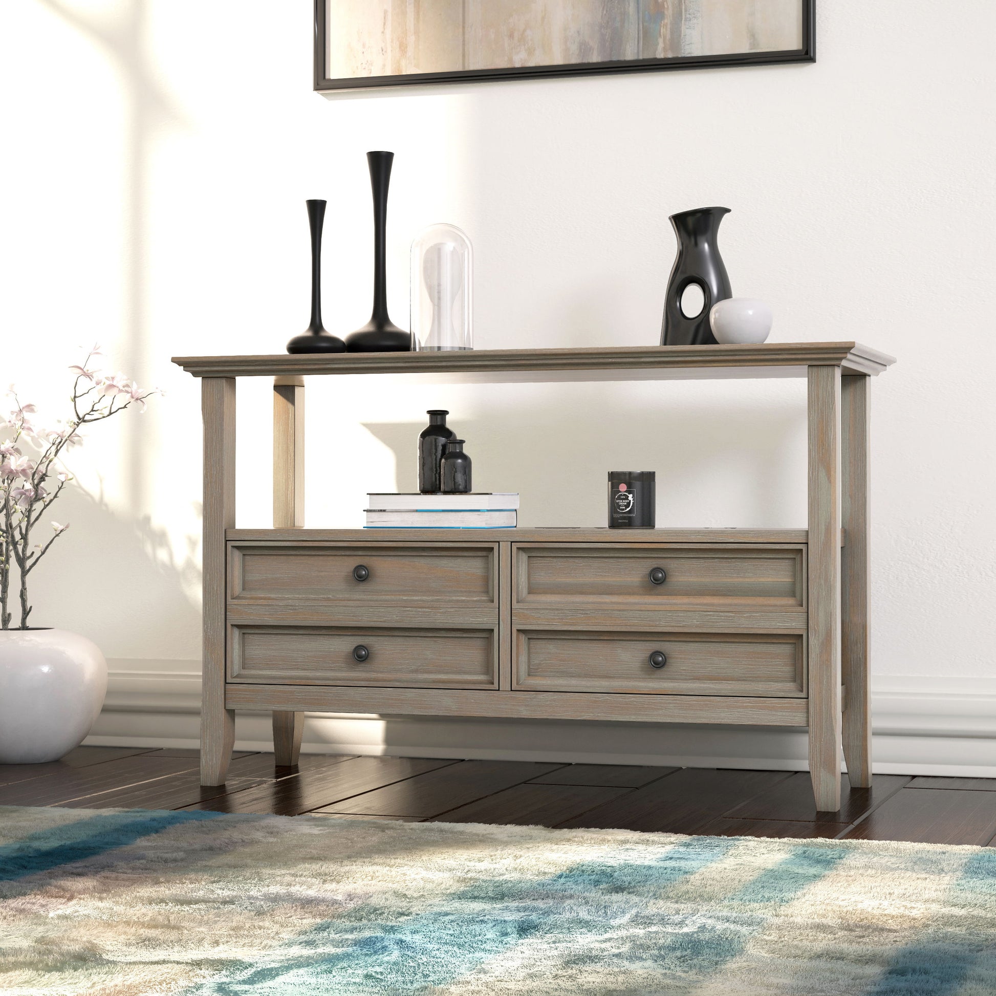 The Amherst Handcrafted Console Sofa Table features four drawers and an open shelf. It’s styled with black and clear vases, a small sculpture, a white vase with flowers, and sits near a blue-patterned rug on a dark wood floor.