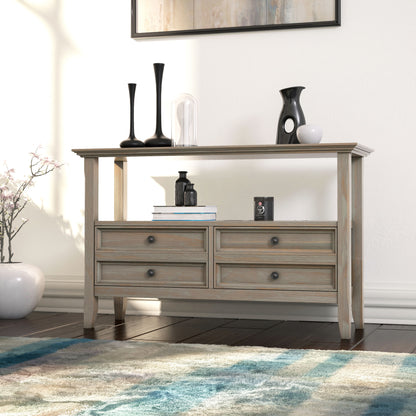 The Amherst Handcrafted Console Sofa Table features four drawers and an open shelf. It’s styled with black and clear vases, a small sculpture, a white vase with flowers, and sits near a blue-patterned rug on a dark wood floor.