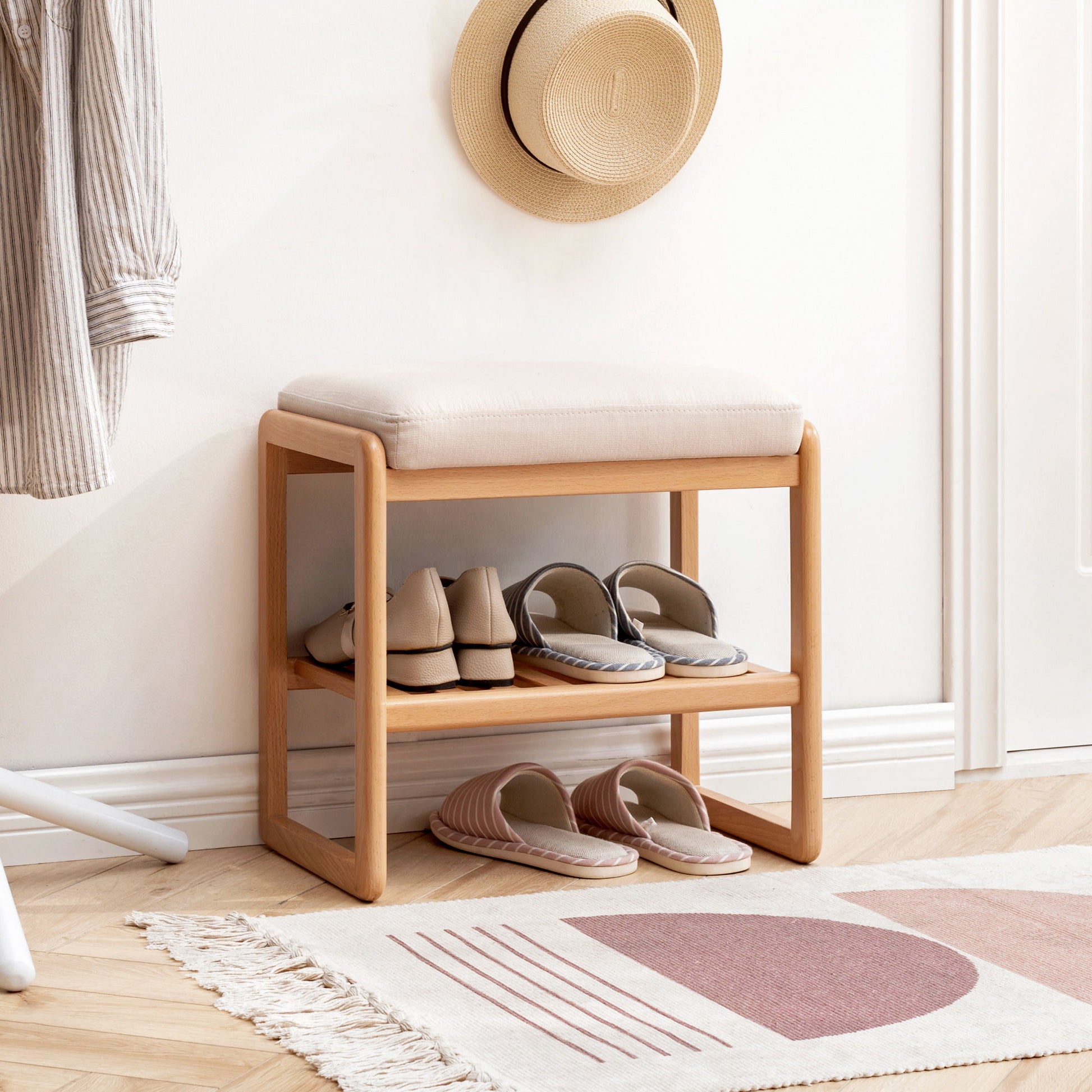 A Shoe Bench, Beech Wood Storage Rack Organizer with a high rebound sponge cushion holds slippers on two shelves below. A straw hat hangs above, a patterned rug is in front on the light wood floor, and a striped shirt hangs on the left.