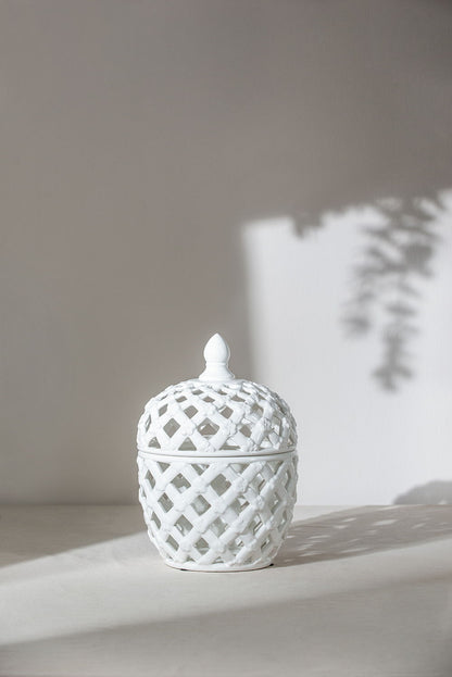 The Ballas - Bird Lidded Jar - White, a ceramic jar with a lattice pattern and pointed lid, sits on a light surface as leafy shadows are cast on the wall behind it.
