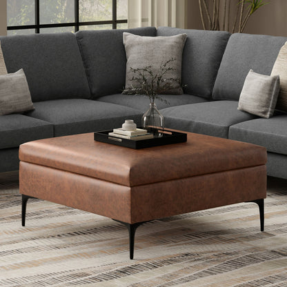 The Rebecca Multifunctional Coffee Table Storage Ottoman with Lift Up Lid sits on a patterned rug in front of a gray sectional sofa, topped with a black tray and glass vase, as light streams in from a window.