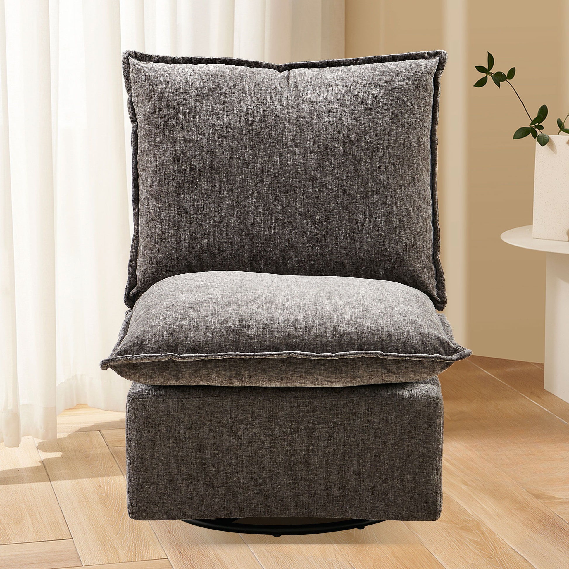 The Detachable Chenille Single 360 Swivel Rocking Rocker, a modern gray armless sofa chair, sits on a light wood floor with sheer white curtains behind and a small white table holding a potted plant to the right.