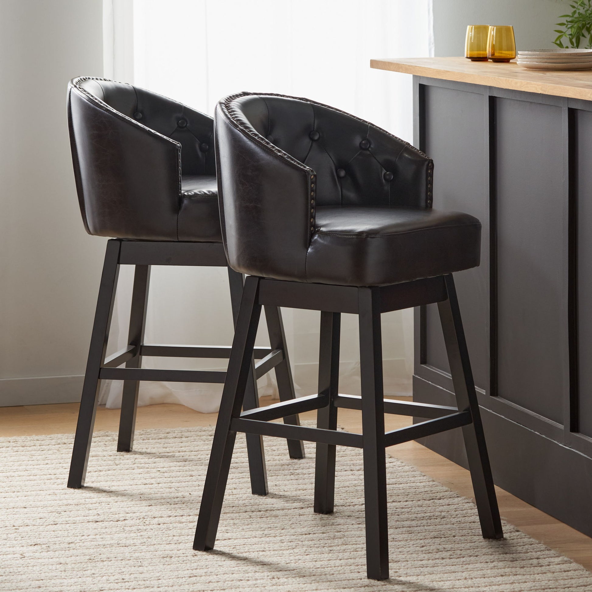 Two Mellifluous 360° swivel bar stools, featuring dark brown leather upholstery, tufted backs, black wooden legs, and elegant nailhead trim, are placed by a kitchen counter in a bright room with light curtains and a neutral rug.