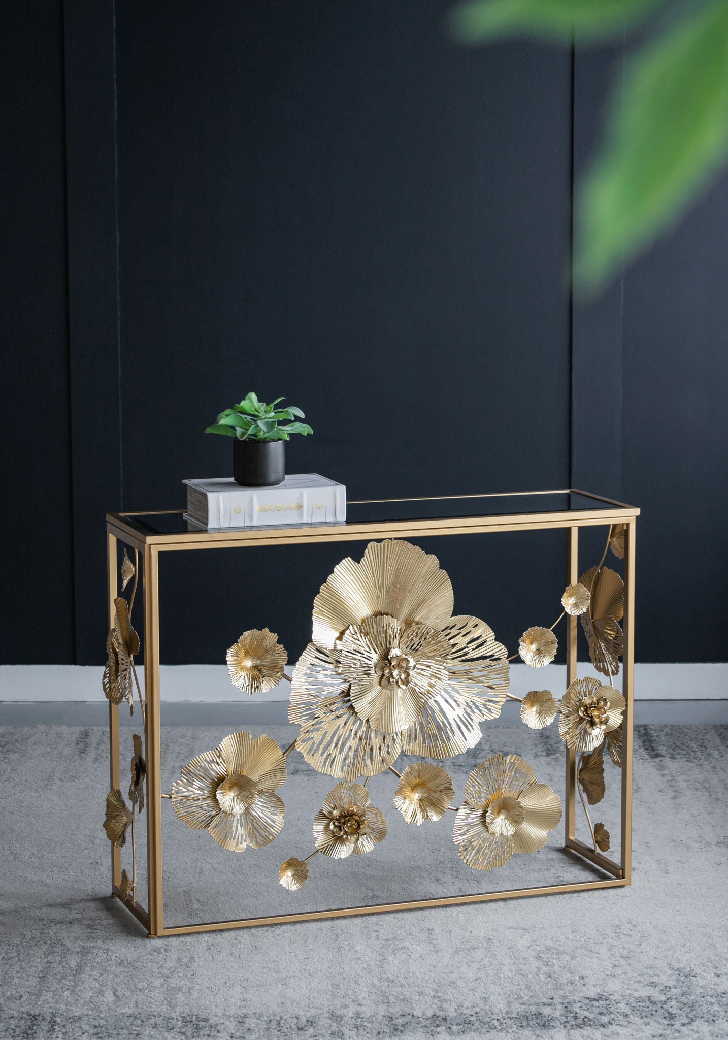A Console Table With Mirrored Glass Top, Modern Entryway Table For Living Room Foyer Entryway - Gold stands against a dark wall, adorned with large gold flowers. A small potted plant and a stack of books rest on the table.